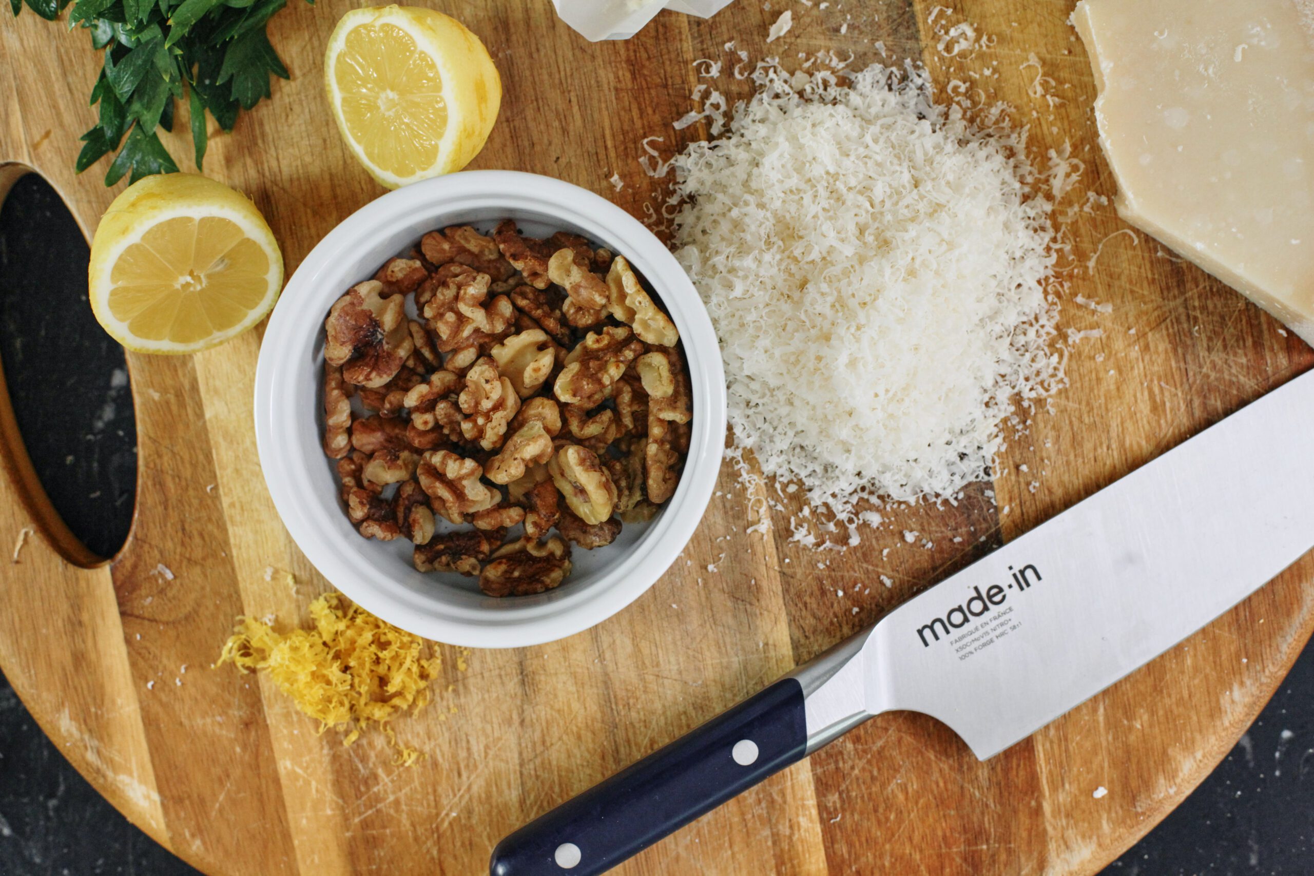 a cutting board with parsley, a halved lemon, lemon zest, a ramekin of walnuts, grated parmesan cheese, and a knife