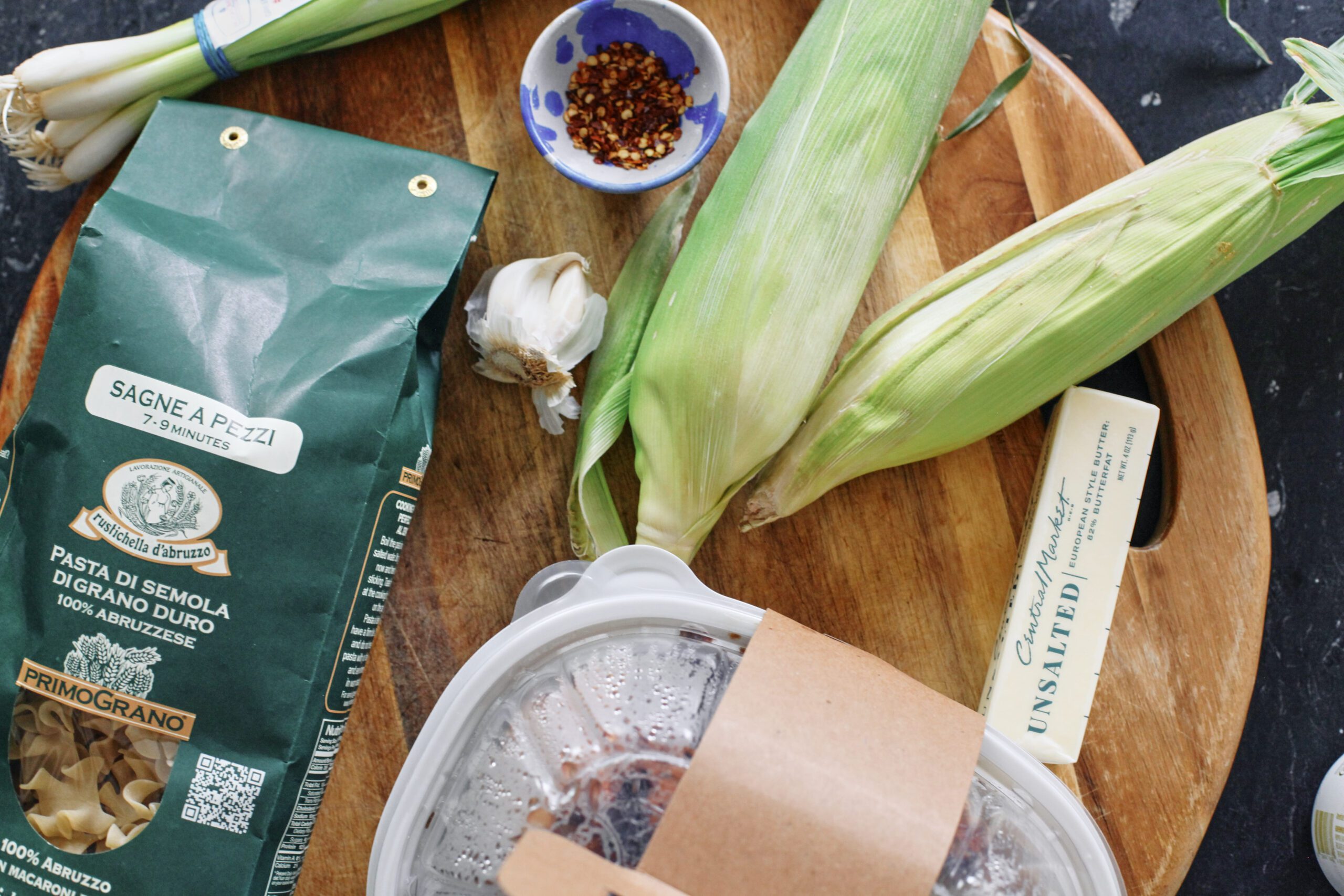 Creamy Corn and Chicken Pasta ingredients for the dish: bag of pasta, ears of corn, bunch of green onions, bulb of garlic, chili flakes in a bowl, rotisserie chicken in a plastic container, and a stick of butter