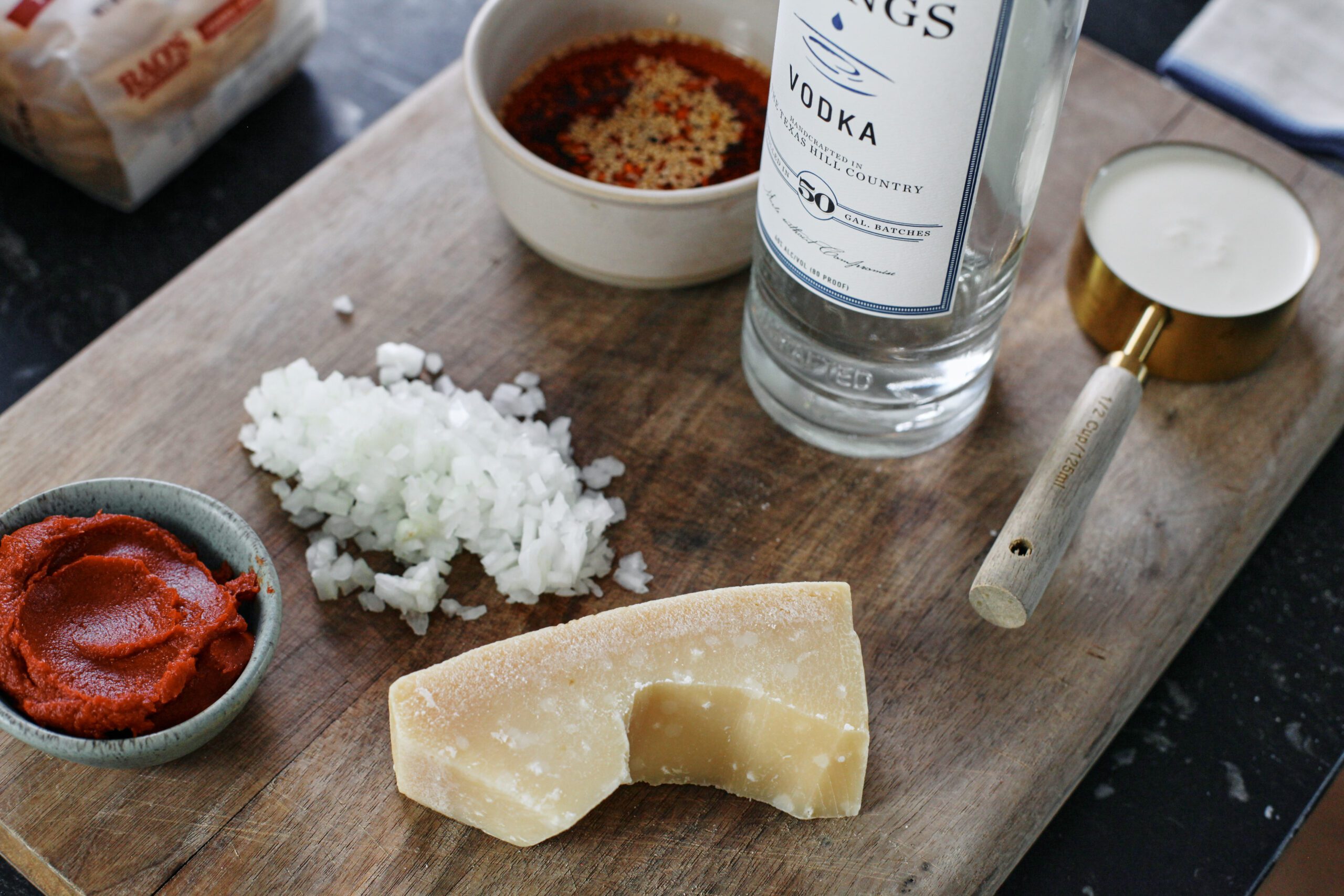 Ingredients on a cutting board prepped for Chili Oil Penne alla Vodka: a bag of pasta, tomato paste in a bowl, chopped onion, a chunk of parmesan cheese, a bowl of chili oil, a bottle of vodka, and heavy cream in a measuring cup.