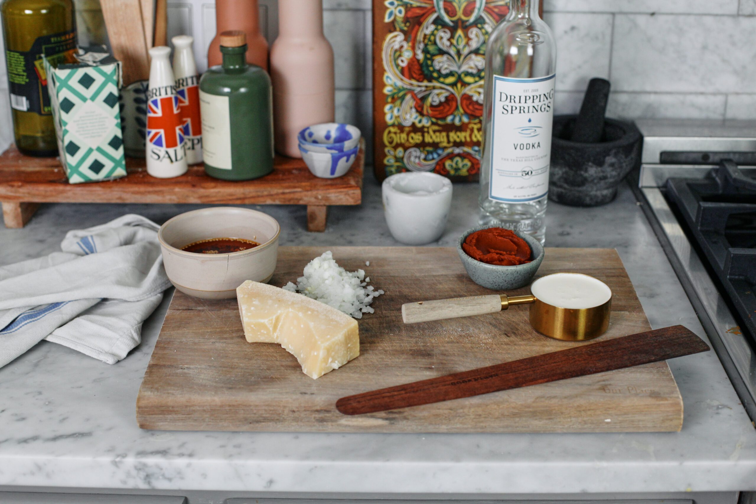 Ingredients on a cutting board prepped for Chili Oil Penne alla Vodka: tomato paste in a bowl, chopped onion, a chunk of parmesan cheese, a bowl of chili oil, a bottle of vodka, and heavy cream in a measuring cup.