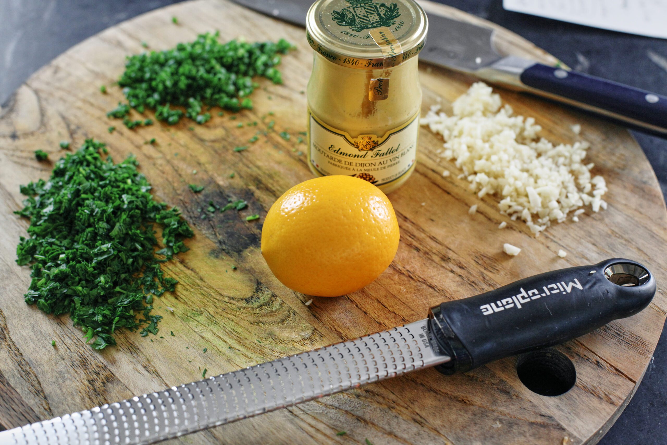 ingredients prepped on a wood cutting board: chopped parsley and green onions, a lemon, a microplane zester, minced garlic, and a jar of dijon mustard