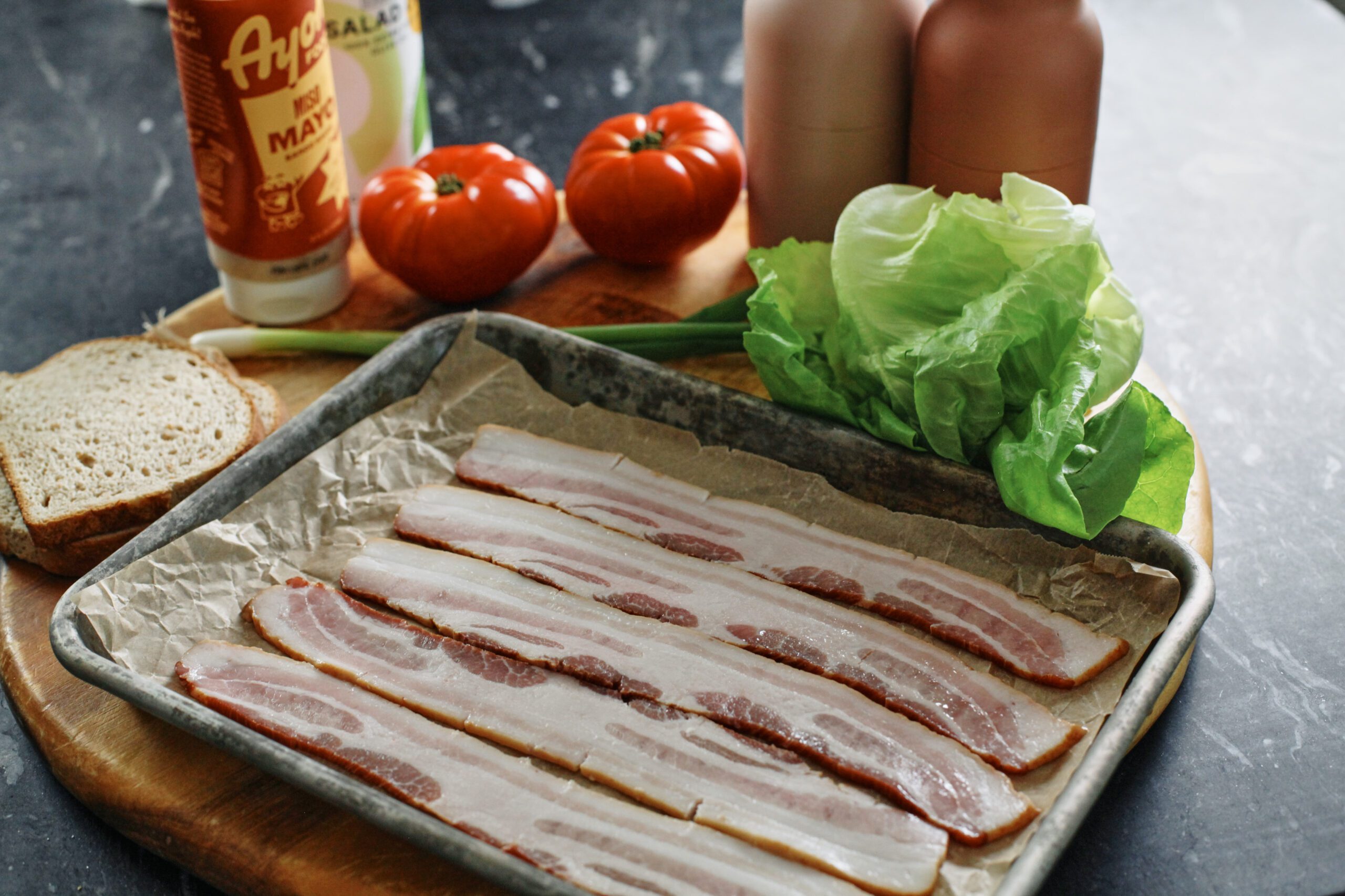 ingredients for BLT: raw bacon on a parchment-lined sheet pan, bread, mayonnaise, green onion, tomatoes, lettuce, salt and pepper mills