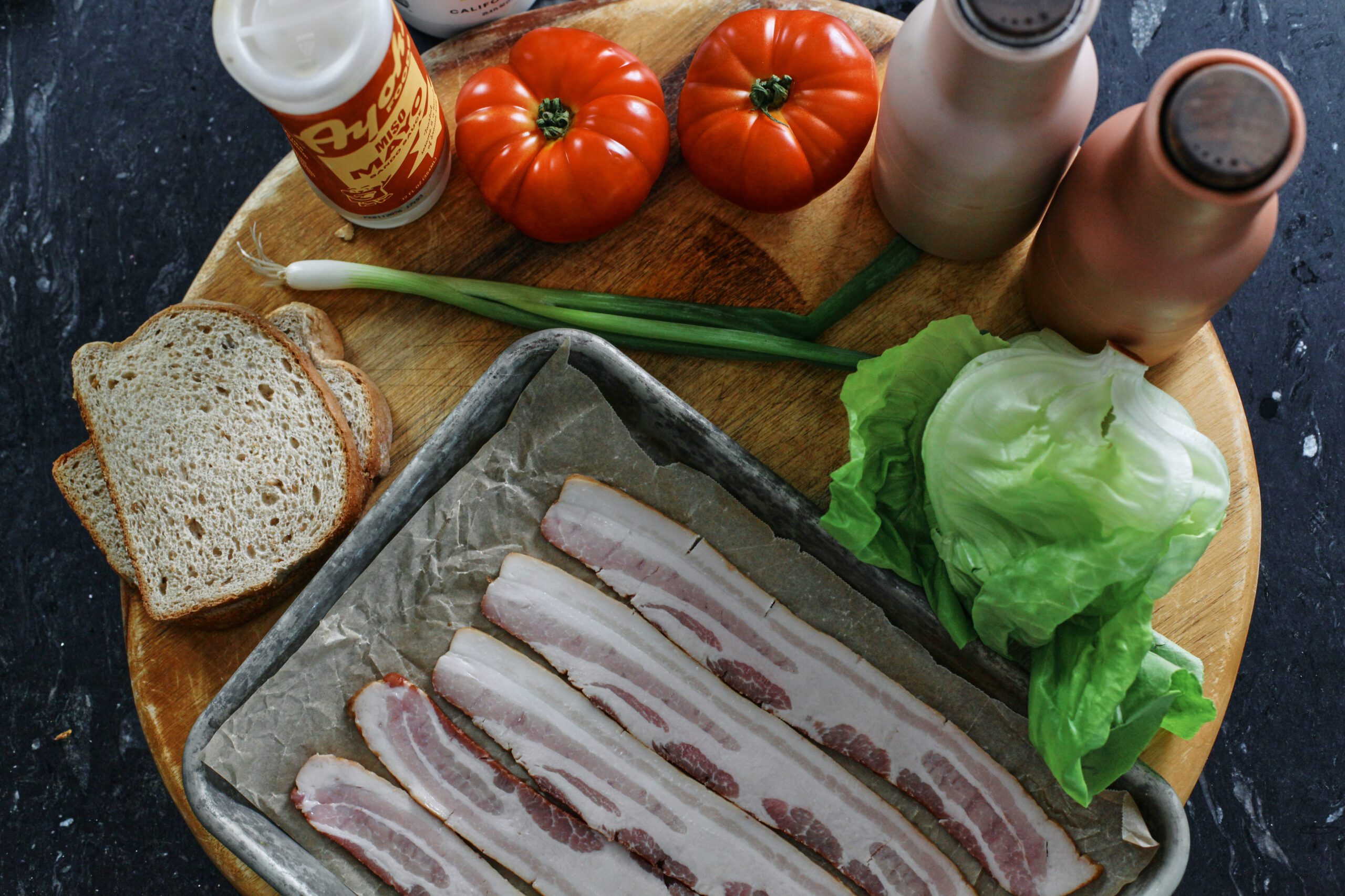 ingredients for BLT: raw bacon on a parchment-lined sheet pan, bread, mayonnaise, green onion, tomatoes, lettuce, salt and pepper mills