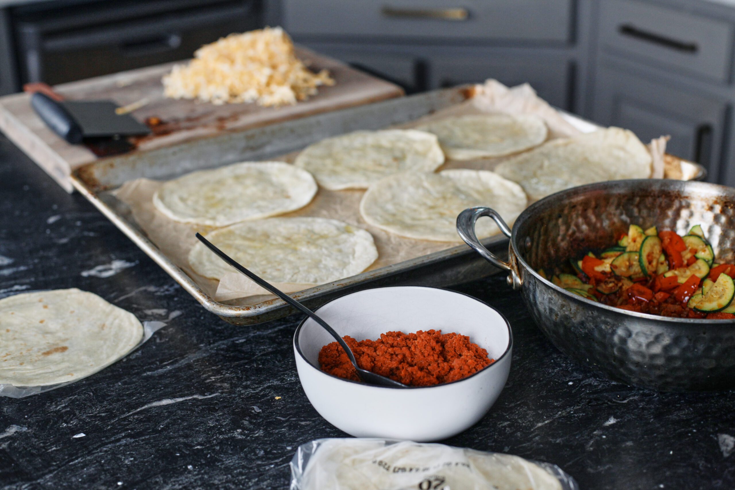 a white bowl of cooked chorizo and a skillet of cooked bell peppers and zucchini. In the background there's a pan of tortillas and shredded cheese.