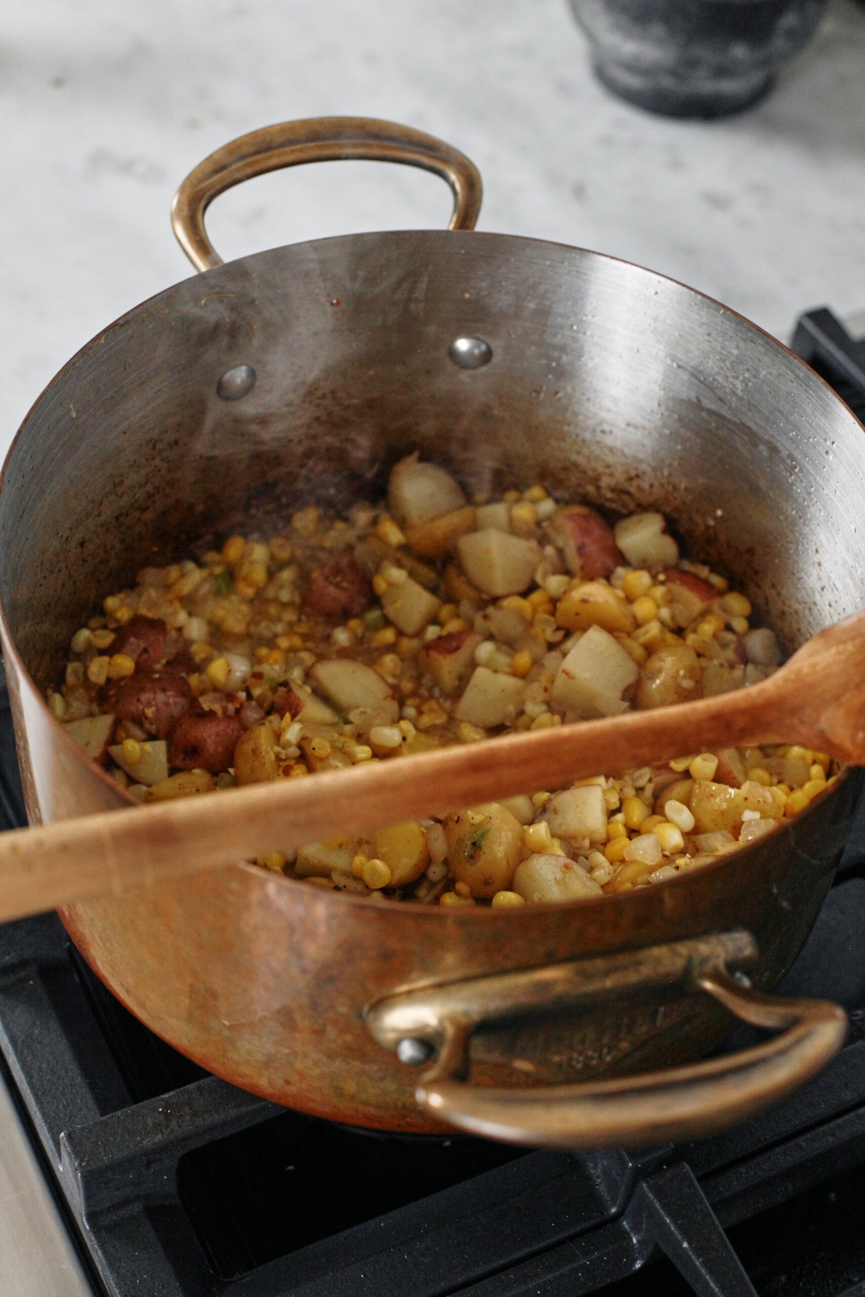 Corn, potatoes, onion, green onion, and minced garlic sautéing in a large metal pot with a wooden spoon resting on the pot.