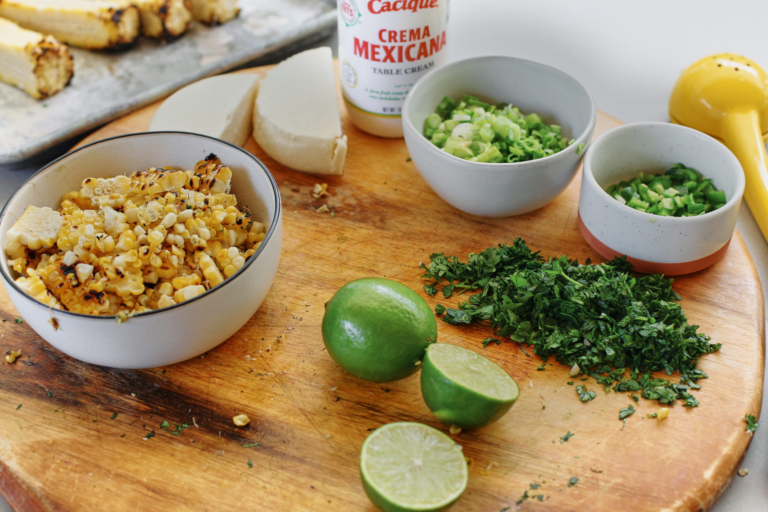 Ingredients prepped/chopped for elote pasta salad: queso fresco, crema mexicana, roasted corn, green onions, jalapeno, cilantro, limes