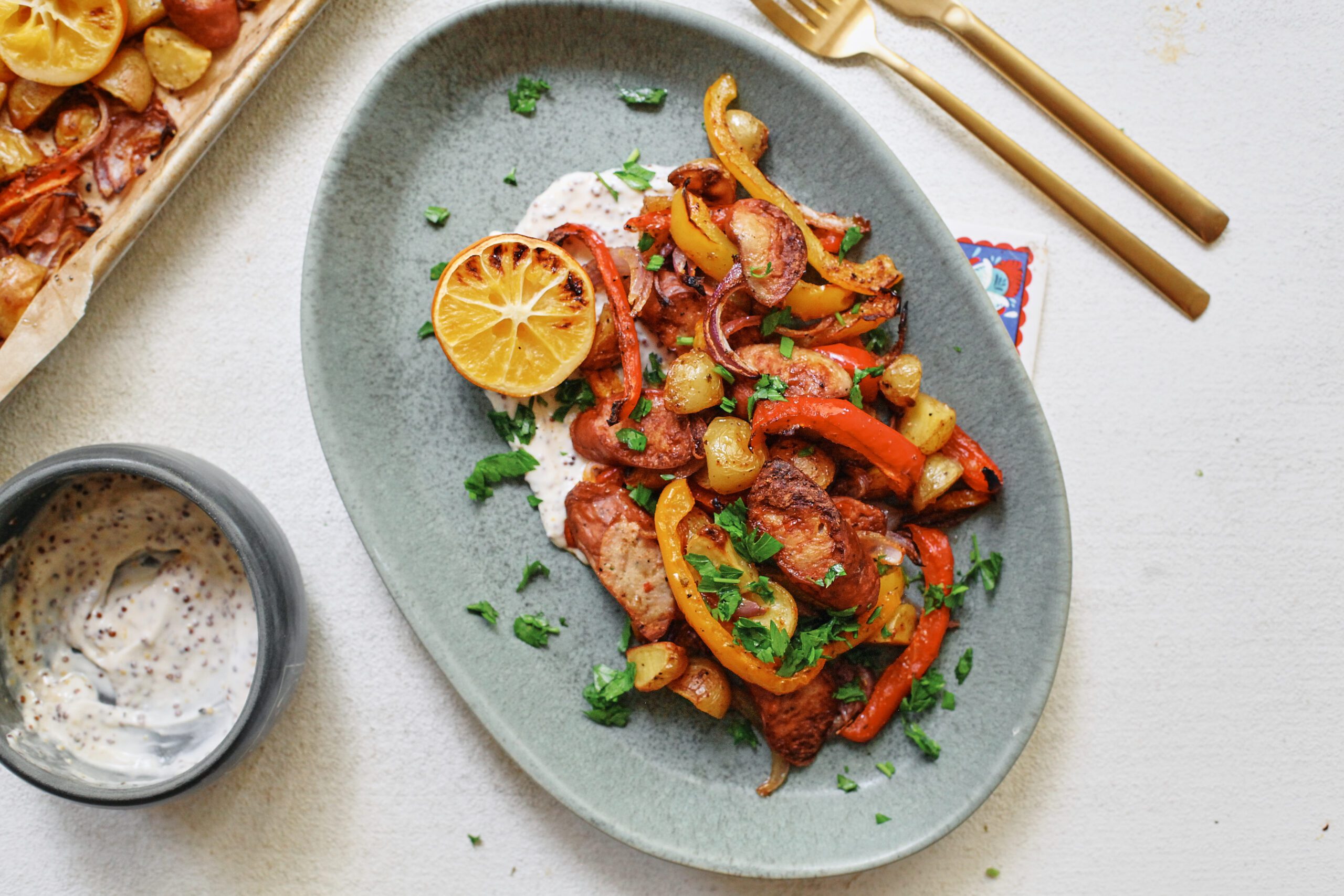 sheet pan sausage, peppers, and potatoes served on a blue plate over a white sauce with half a roasted lemon