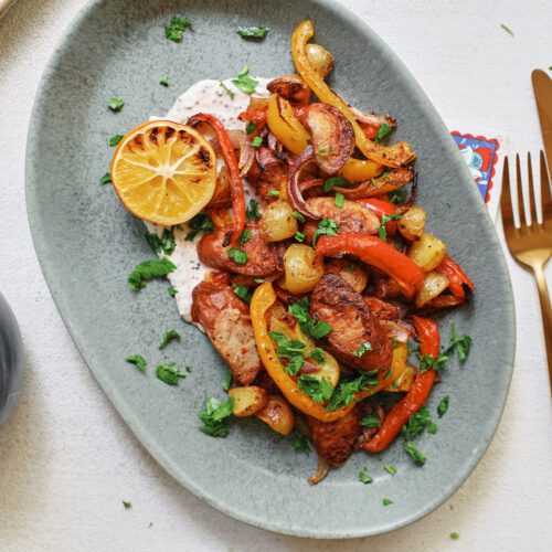sheet pan sausage, peppers, and potatoes served on a blue plate over a white sauce with half a roasted lemon