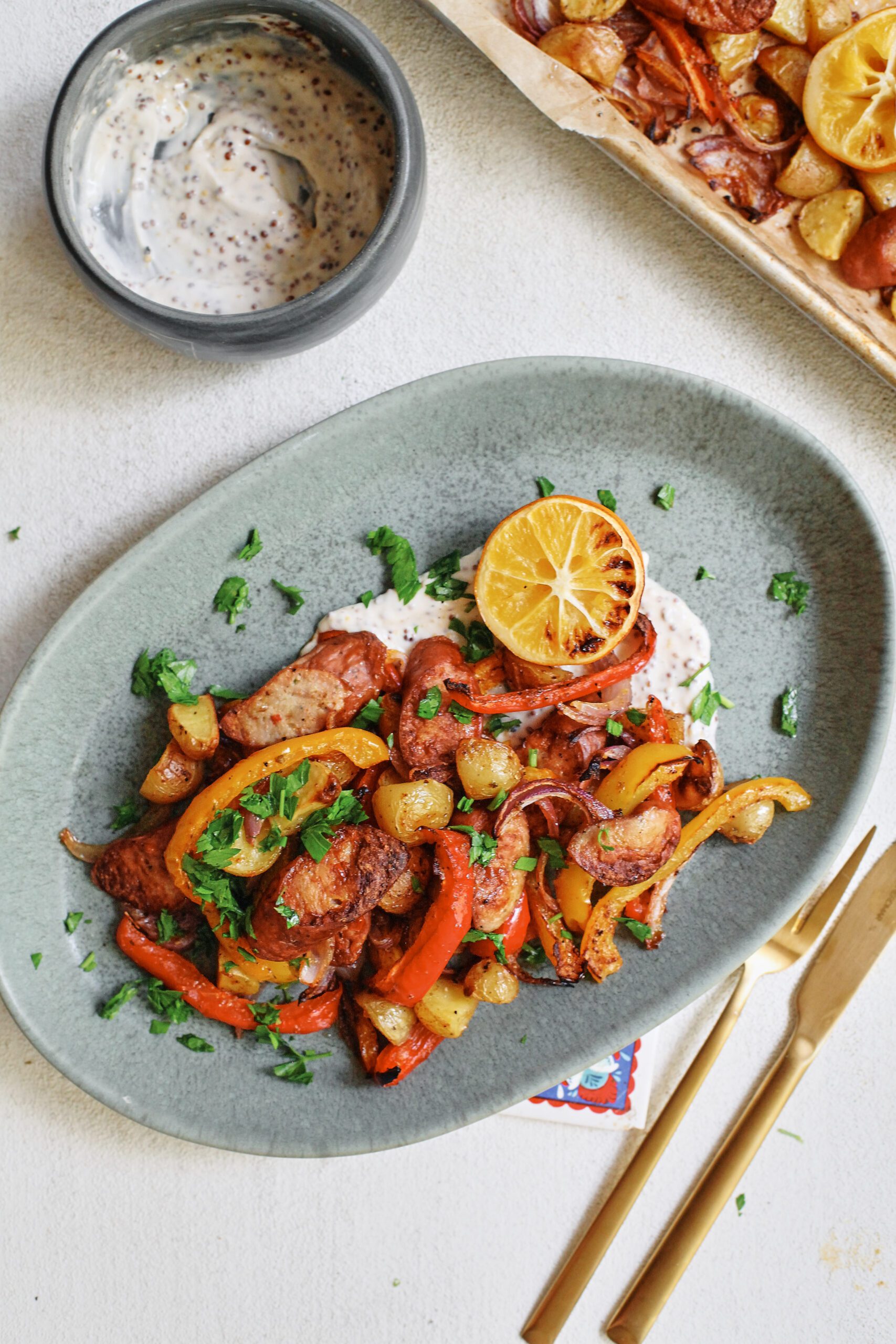 sheet pan sausage, peppers, and potatoes served on a blue plate over a white sauce with half a roasted lemon