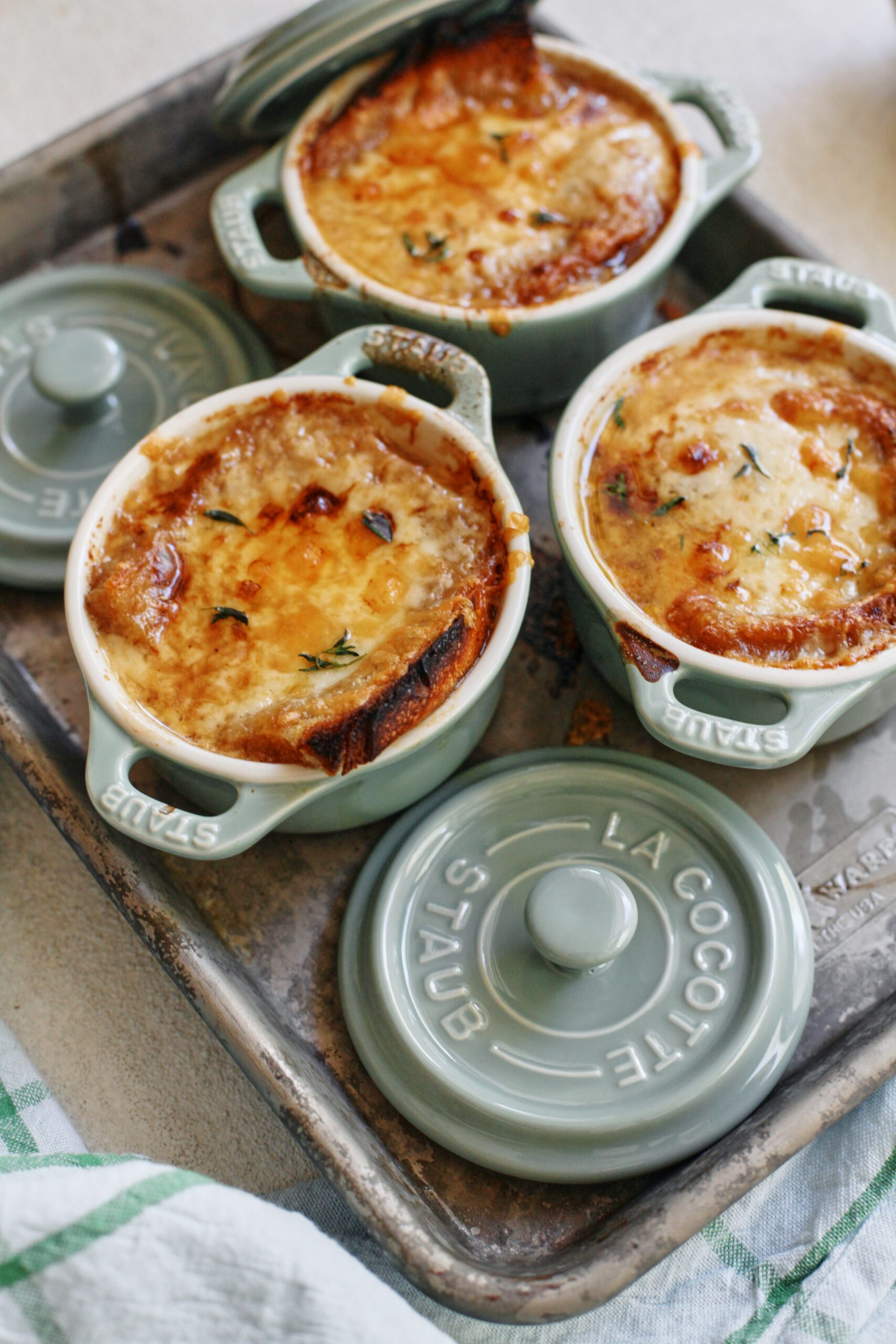 french onion soup in mini green dutch ovens that are arranged on a sheet pan, topped with golden, melted cheese