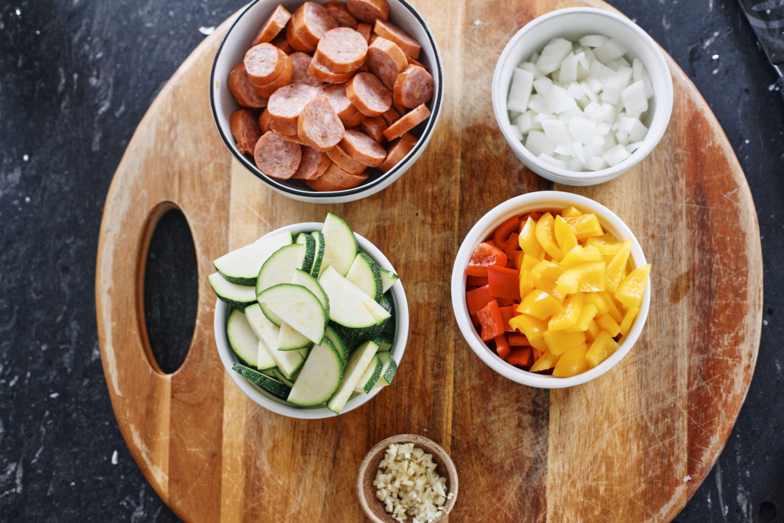 prepped ingredients in bowls: sliced sausages, chopped onion, chopped zucchini, chopped bell peppers, minced garlic