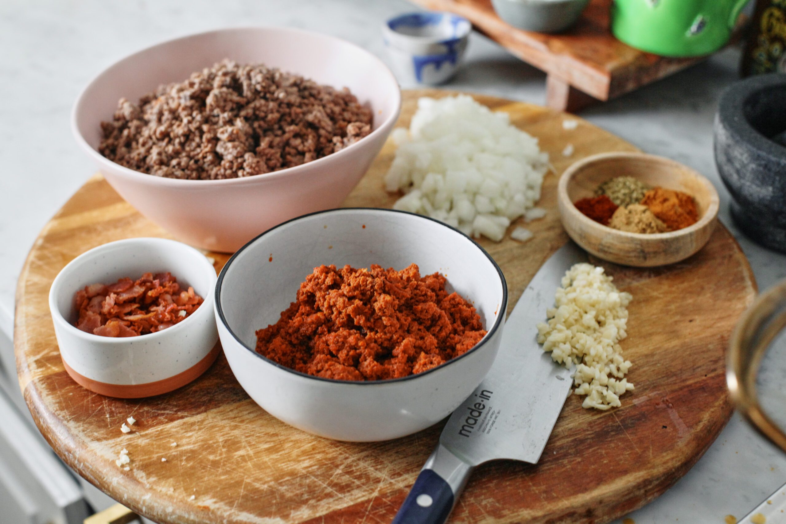 prepped ingredients: bowls of ground beef, chorizo, and chopped bacon with minced garlic, a chopped onion, and a small bowl of spices