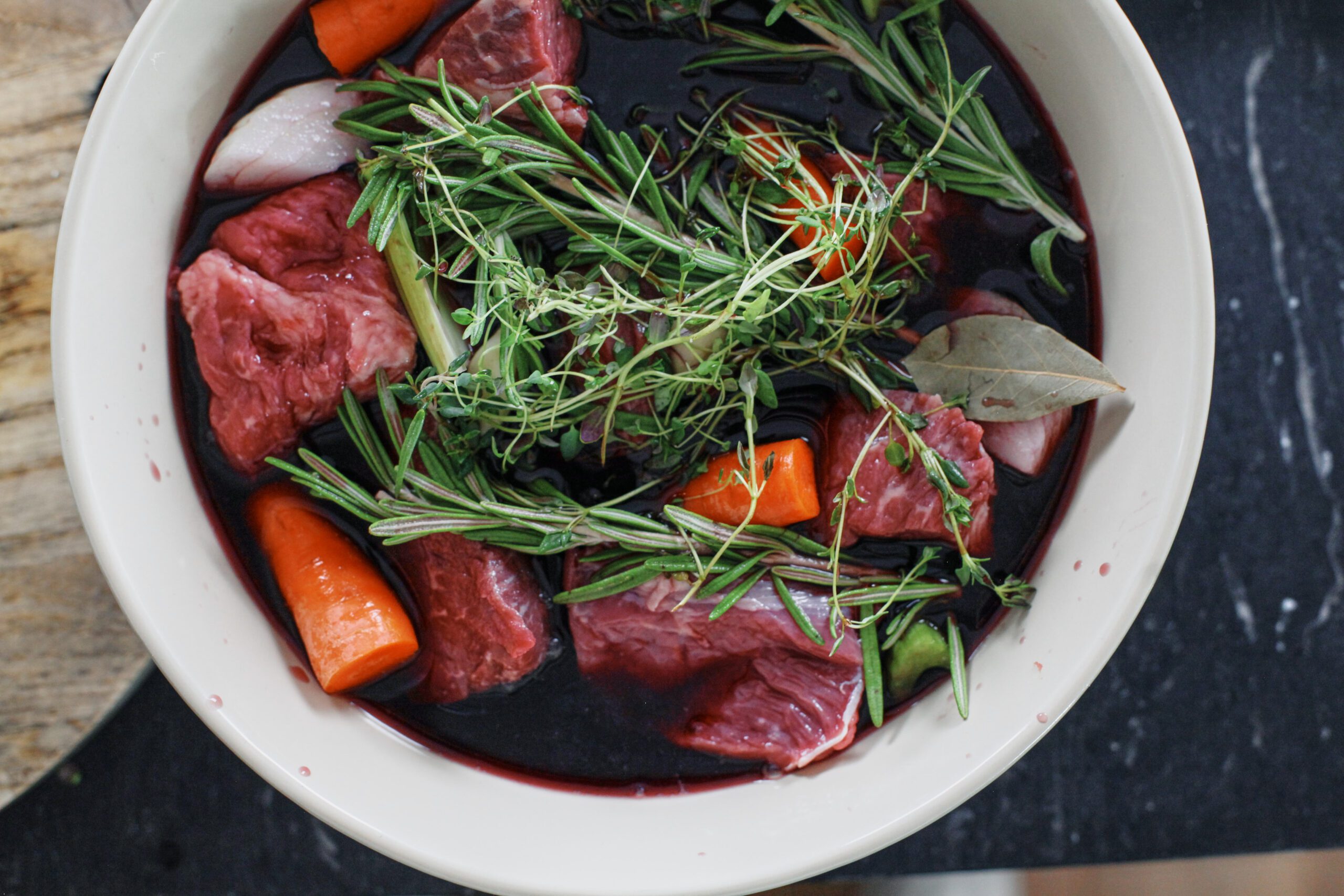 beef in a bowl with the marinade: red wine, thyme, rosemary, a bay leaf, carrots, onion, and celery, garlic cloves
