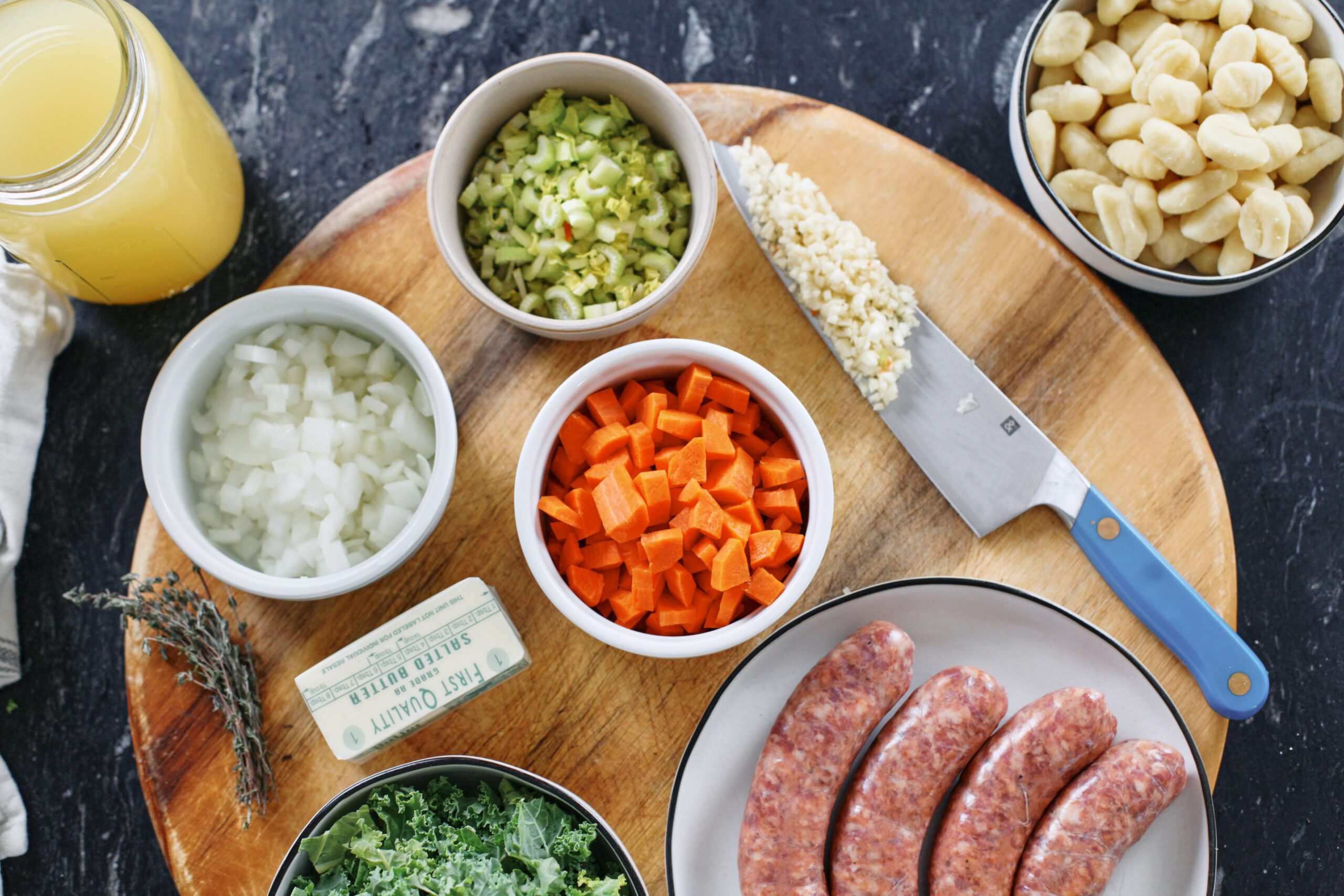 Creamy gnocchi soup with sausage 3 prepped ingredients on a wood cutting board: sausages, chopped carrots, celery, onion, and kale, 1/2 stick of butter, thyme, and garlic on a blue-handled chef's knife
