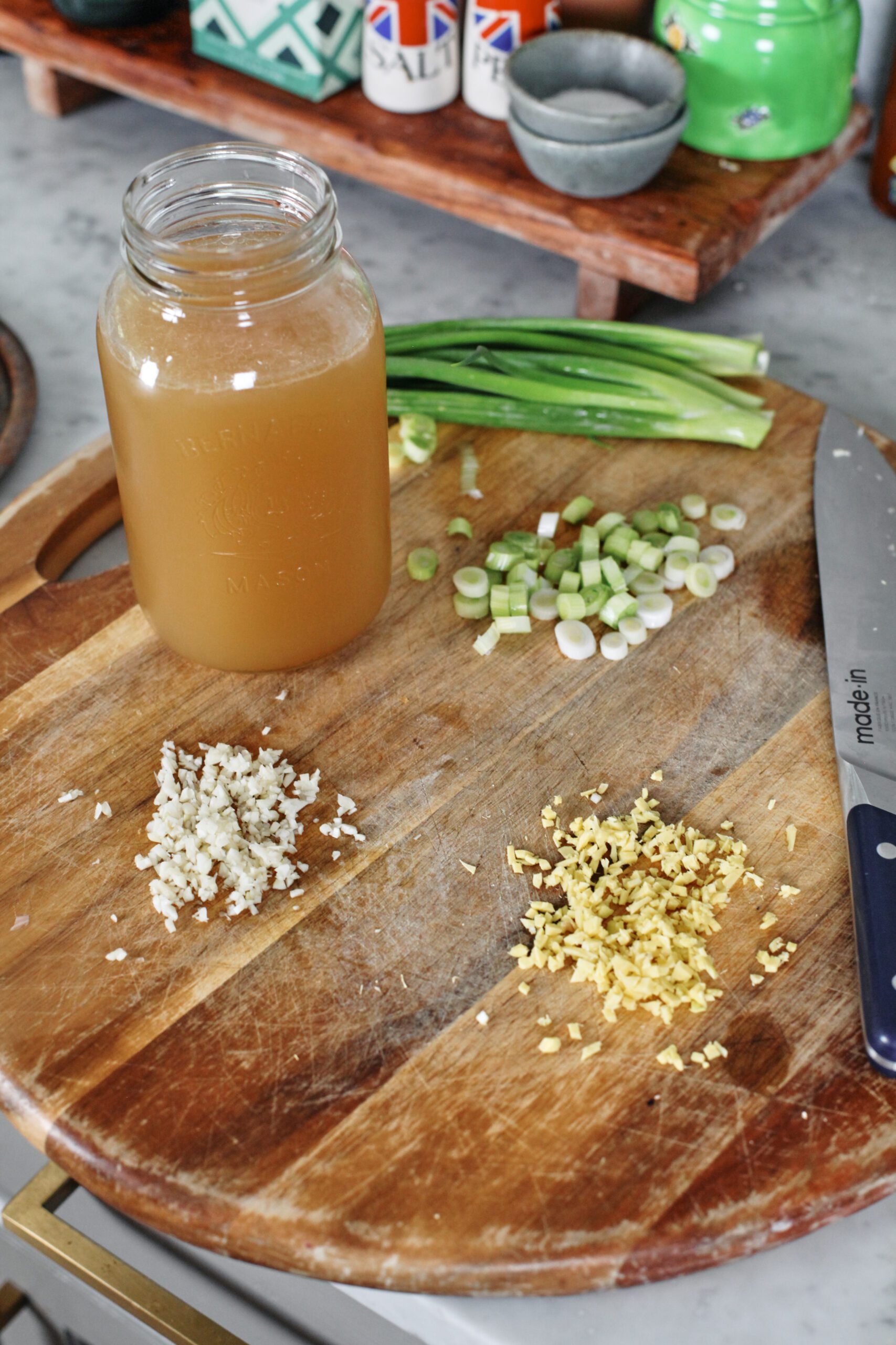a jar of chicken stock on a wood cutting board with minced garlic, minced ginger, and chopped green onions