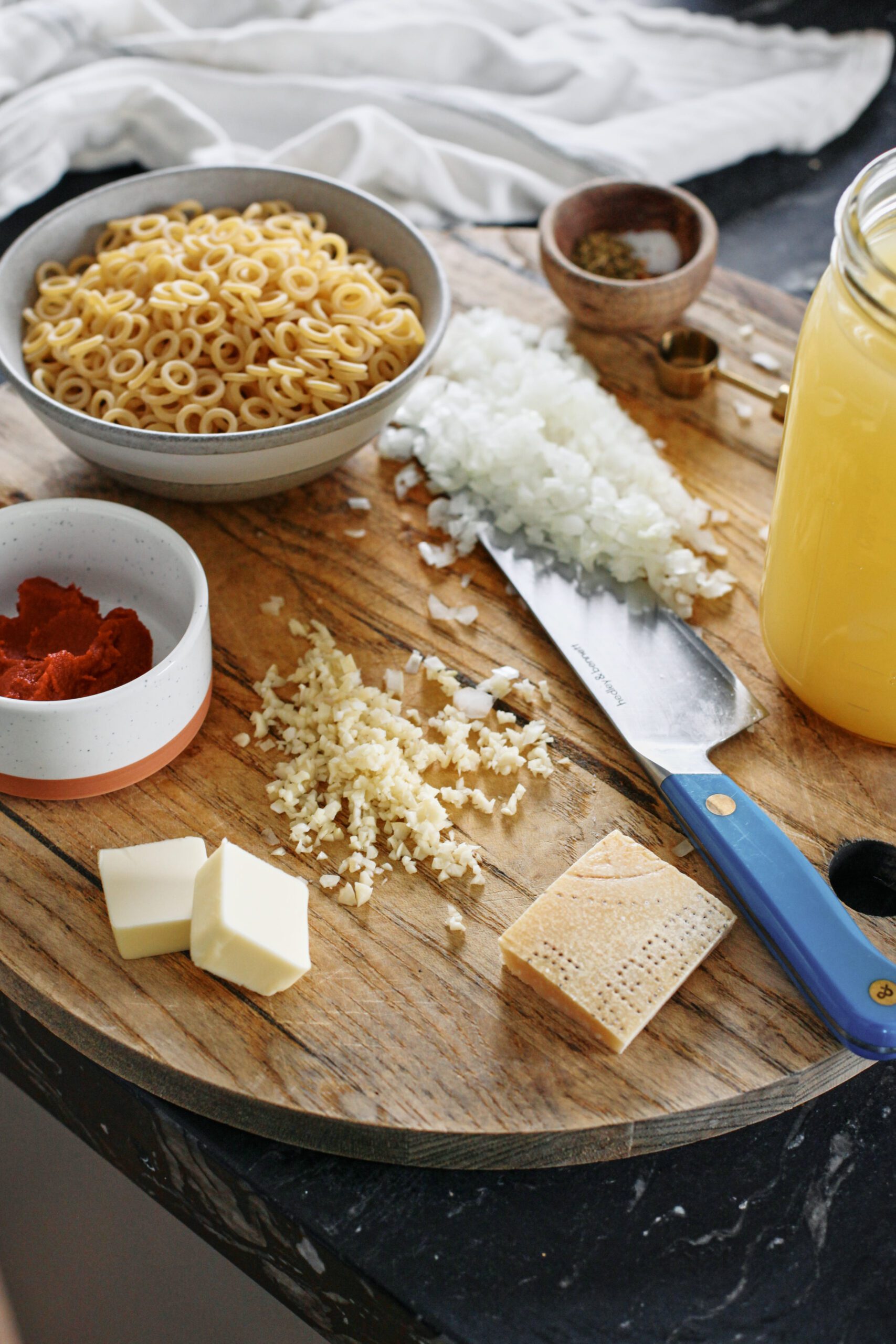 prepped ingredients on a wood cutting board with a blue-handled knife: tomato paste, anelli siciliani pasta, chopped onion, minced garlic, parmesan rind, a small bowl of spices, a jar of chicken stock