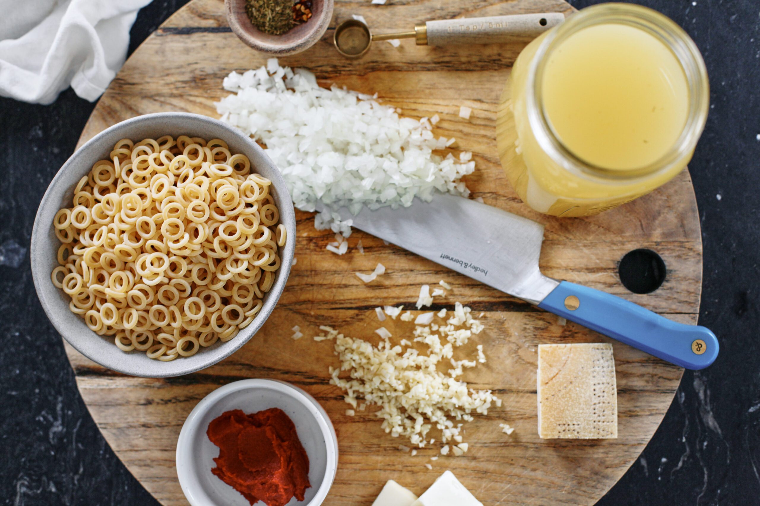 prepped ingredients on a wood cutting board with a blue-handled knife: tomato paste, anelli siciliani pasta, chopped onion, minced garlic, parmesan rind, a small bowl of spices, a jar of chicken stock