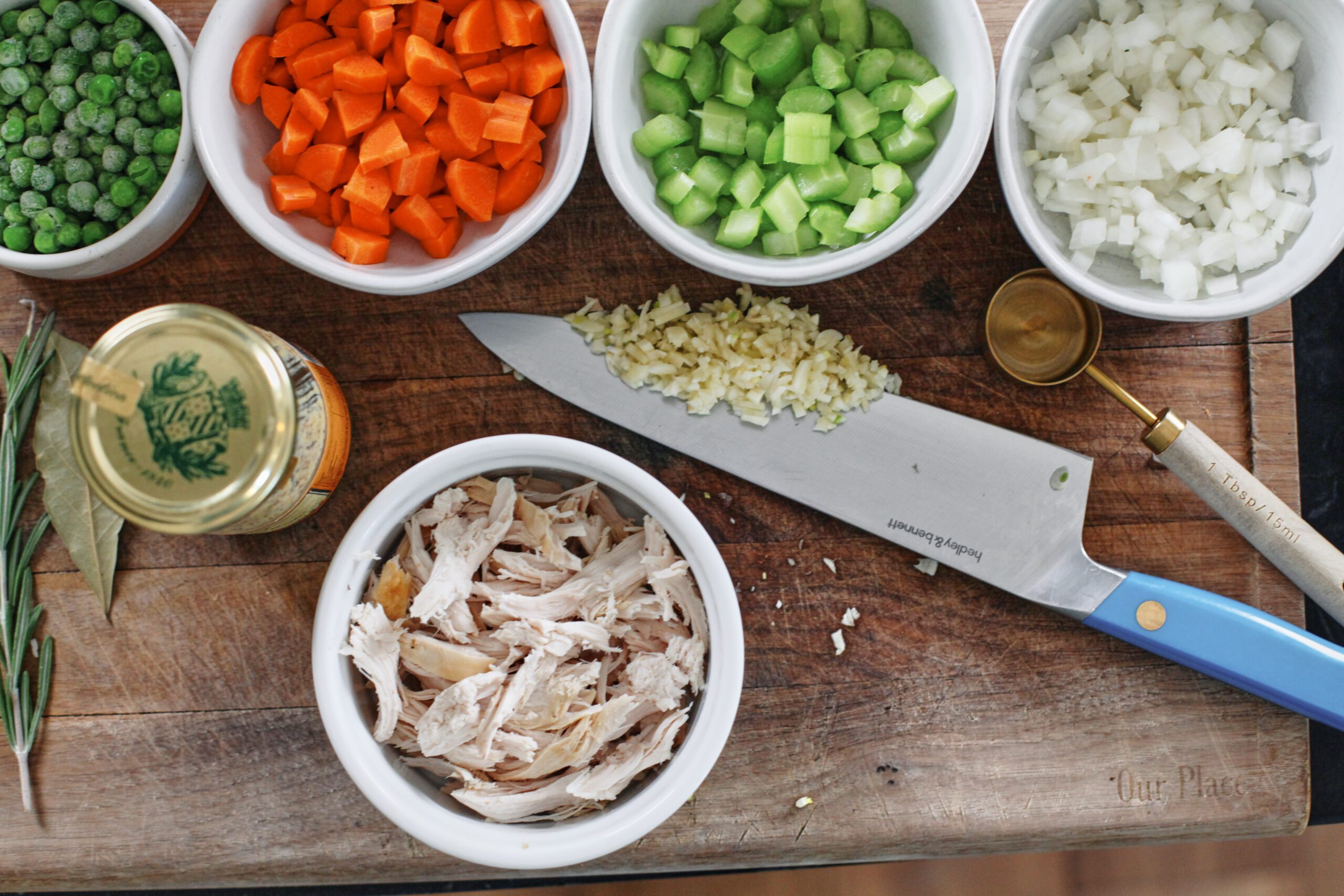 ingredients prepped on a wooden cutting board: rosemary sprig, bay leaf, frozen peas, chopped carrots, chopped celery, chopped onion, minced garlic on a knife, shredded rotisserie chicken, and a jar of mustard