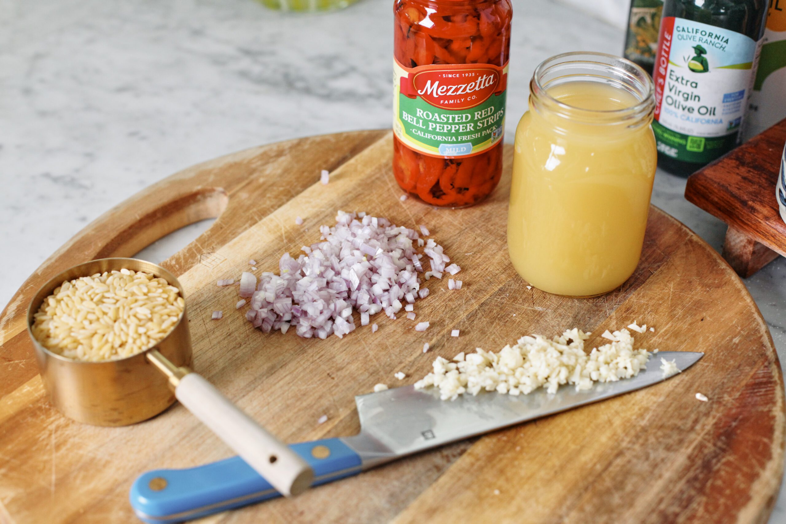ingredients prepped on a wood cutting board: a cup of dry orzo, minced shallot, minced garlic on a chef's knife, a jar of roasted red peppers, a mason jar of broth