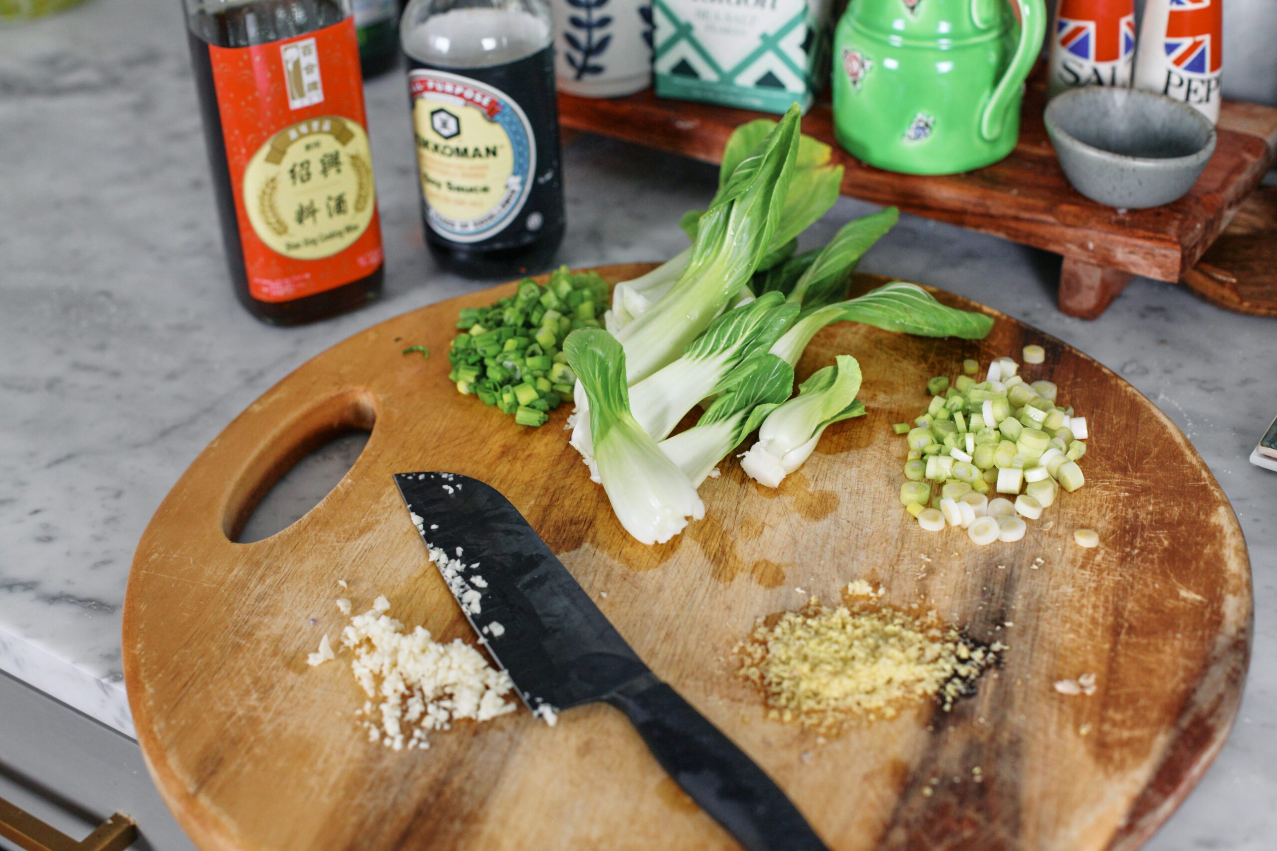 prepped ingredients on a wood cutting board with a knife: baby bok choy, minced garlic, minced ginger, chopped green onion