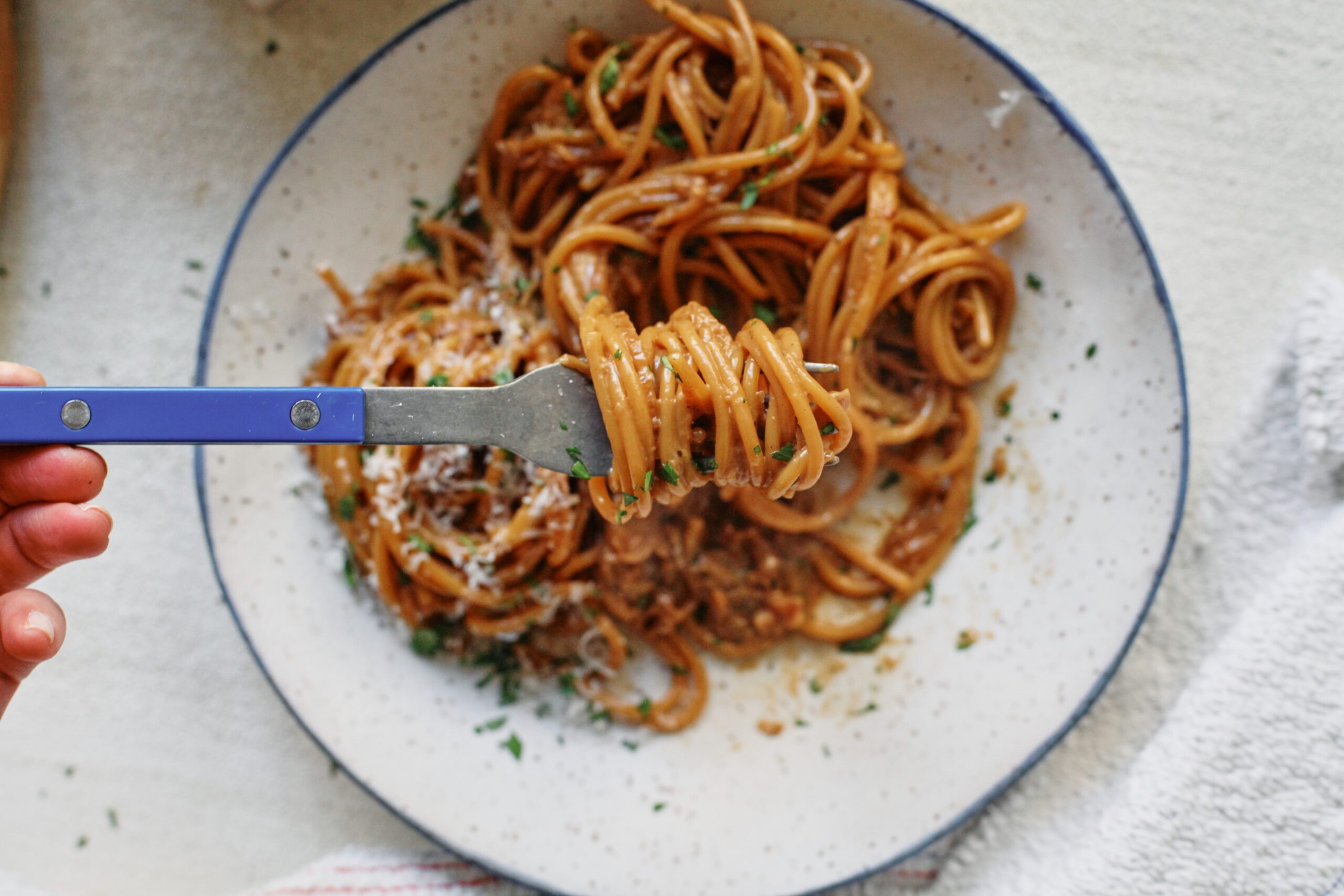A blue-handled fork twirling french onion spaghetti in a shallow white bowl with a black rim