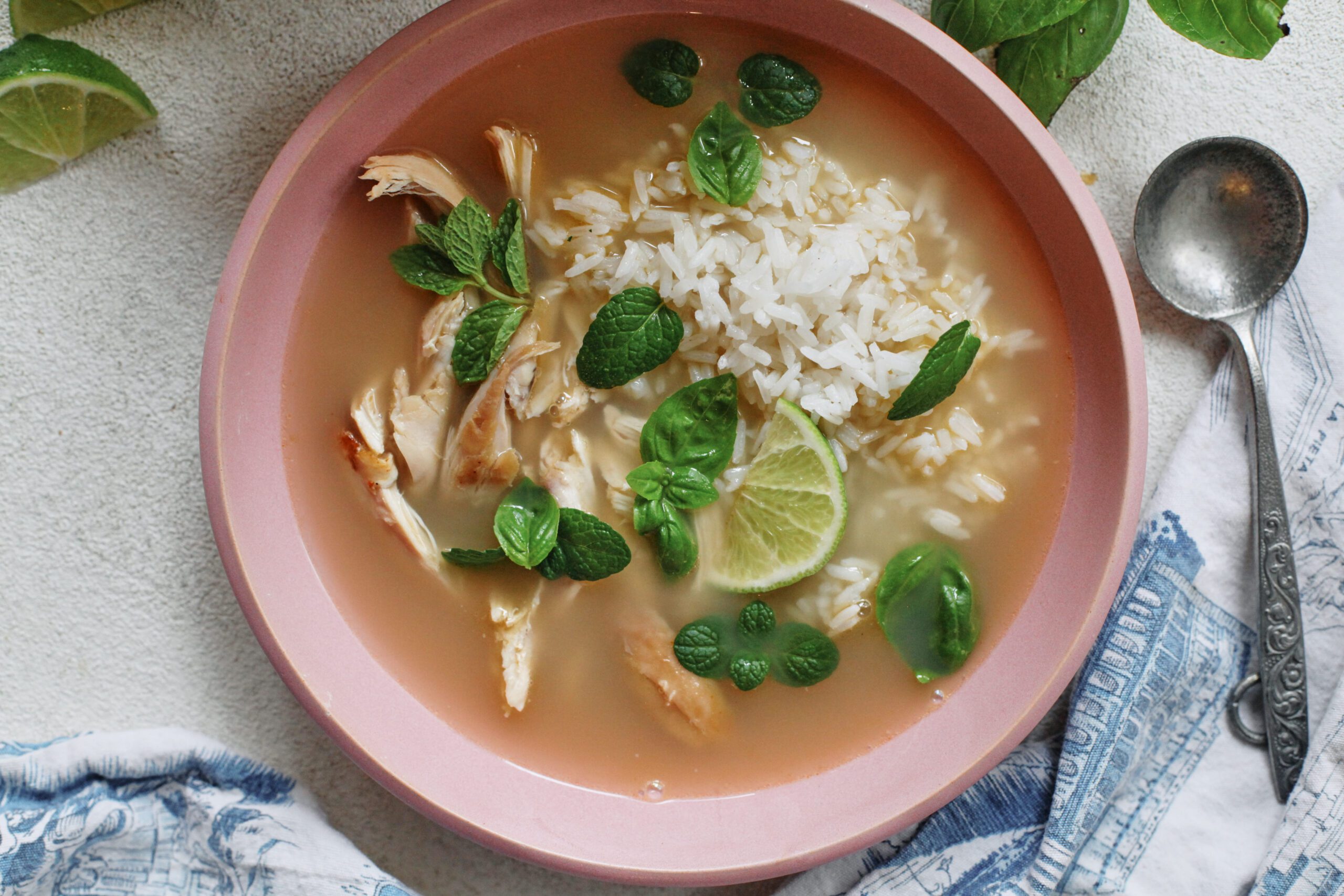 Overhead photo of a bowl of ginger chicken rice soup with shredded rotisserie chicken, fluffy white rice, and fresh mint and basil on top, garnished with a lime wedge