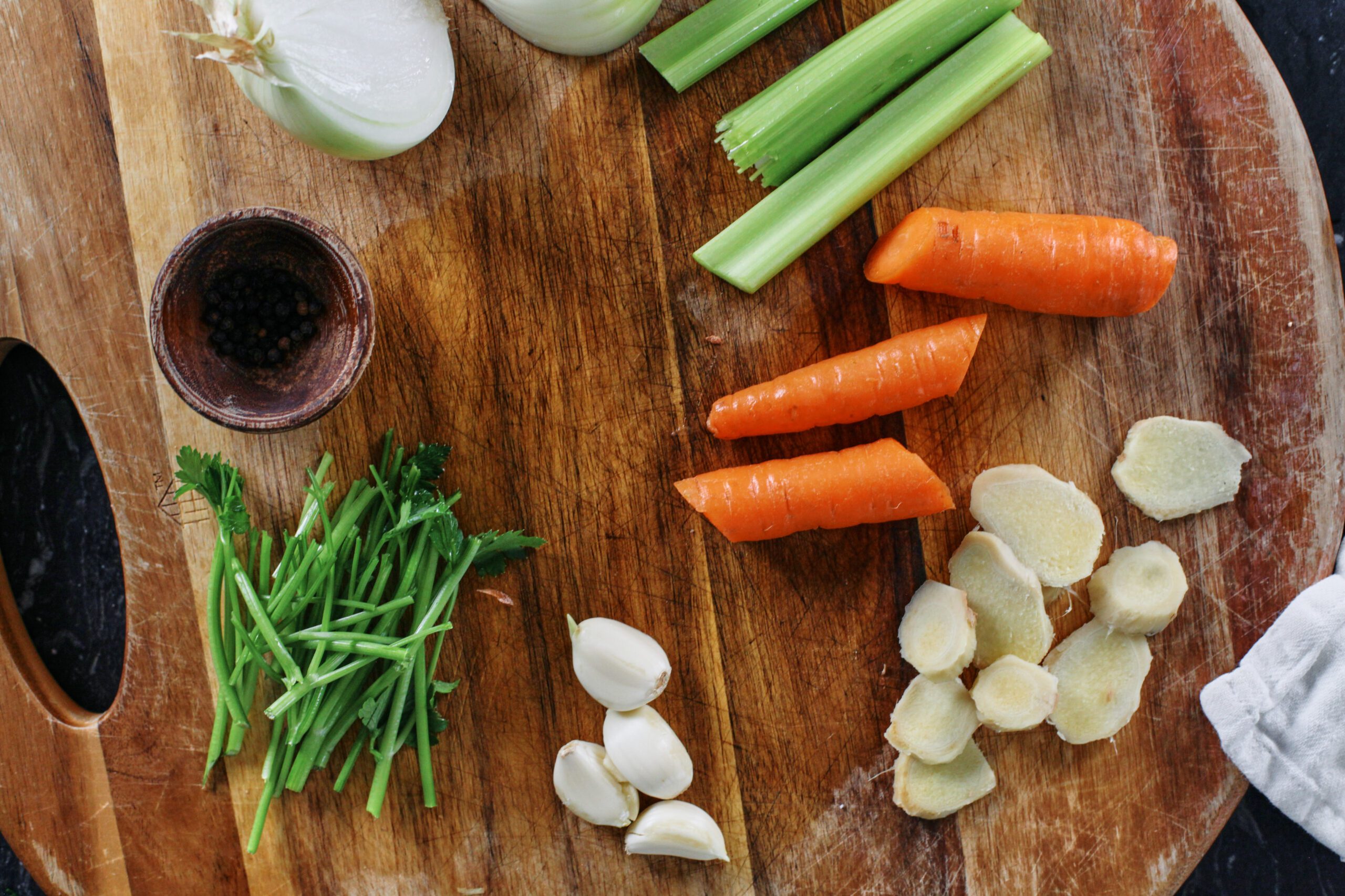 prepped ingredients on a wooden cutting board: a small bowl of peppercorns, a halved onion, celery and carrot cut in thirds, sliced ginger, parsley stems, 4 garlic cloves