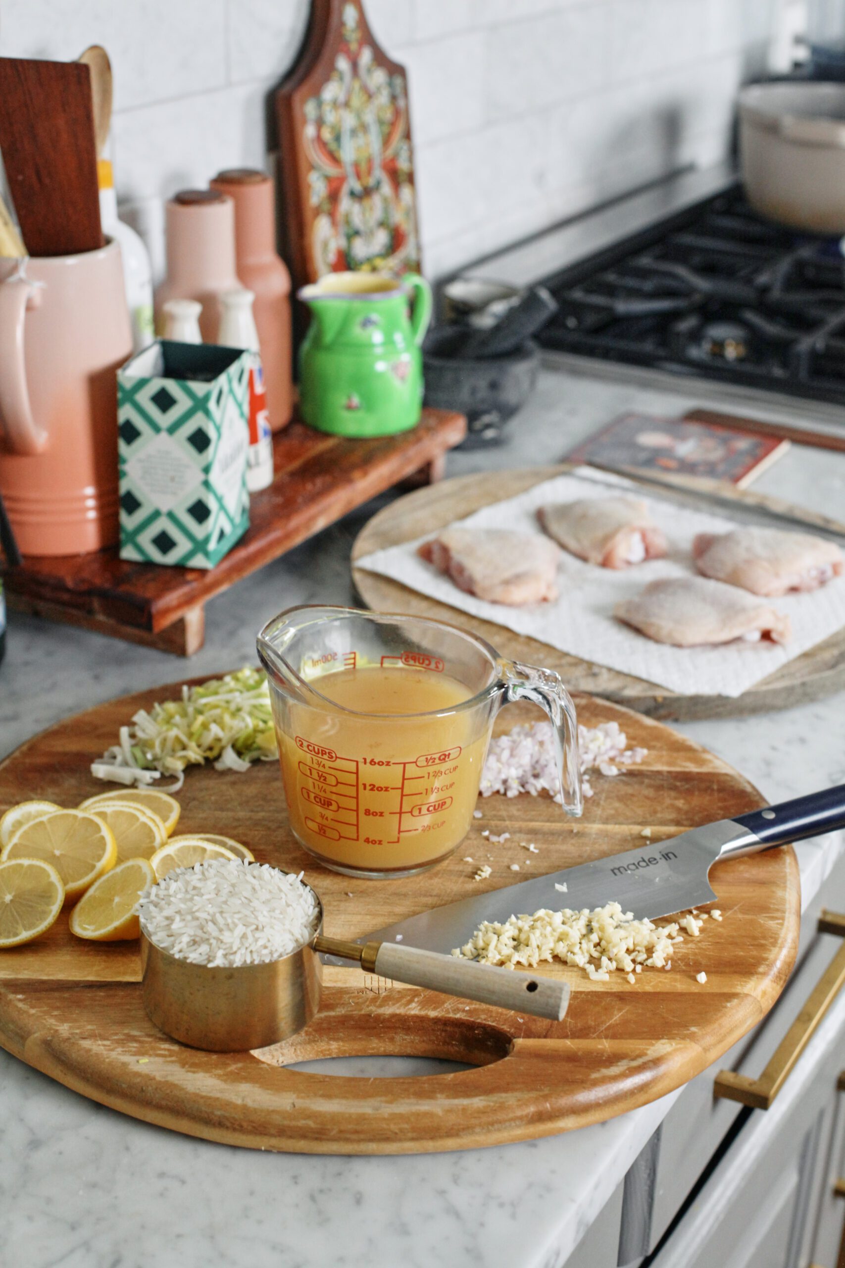 prepped ingredients on a round cutting board: a cup of white rice, lemon slices, minced garlic on a knife, a glass measuring container of broth, a chopped shallot, a minced shallot