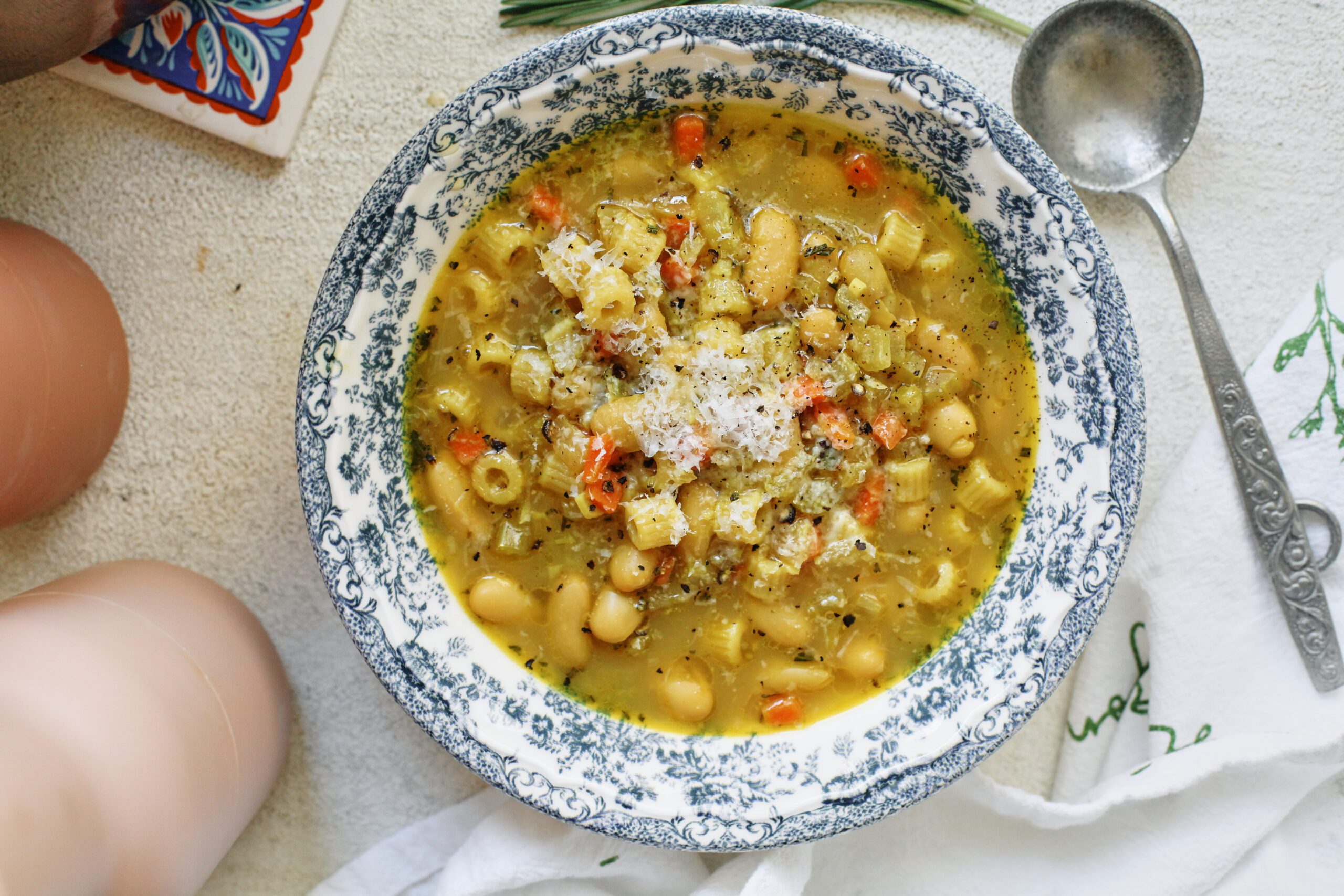 overhead photo of pasta e fagioli alla bianca in a blue/white floral patterned bowl. it's garnished with grated parmesan cheese and black pepper. an antique-looking silver spoon is next to the bowl