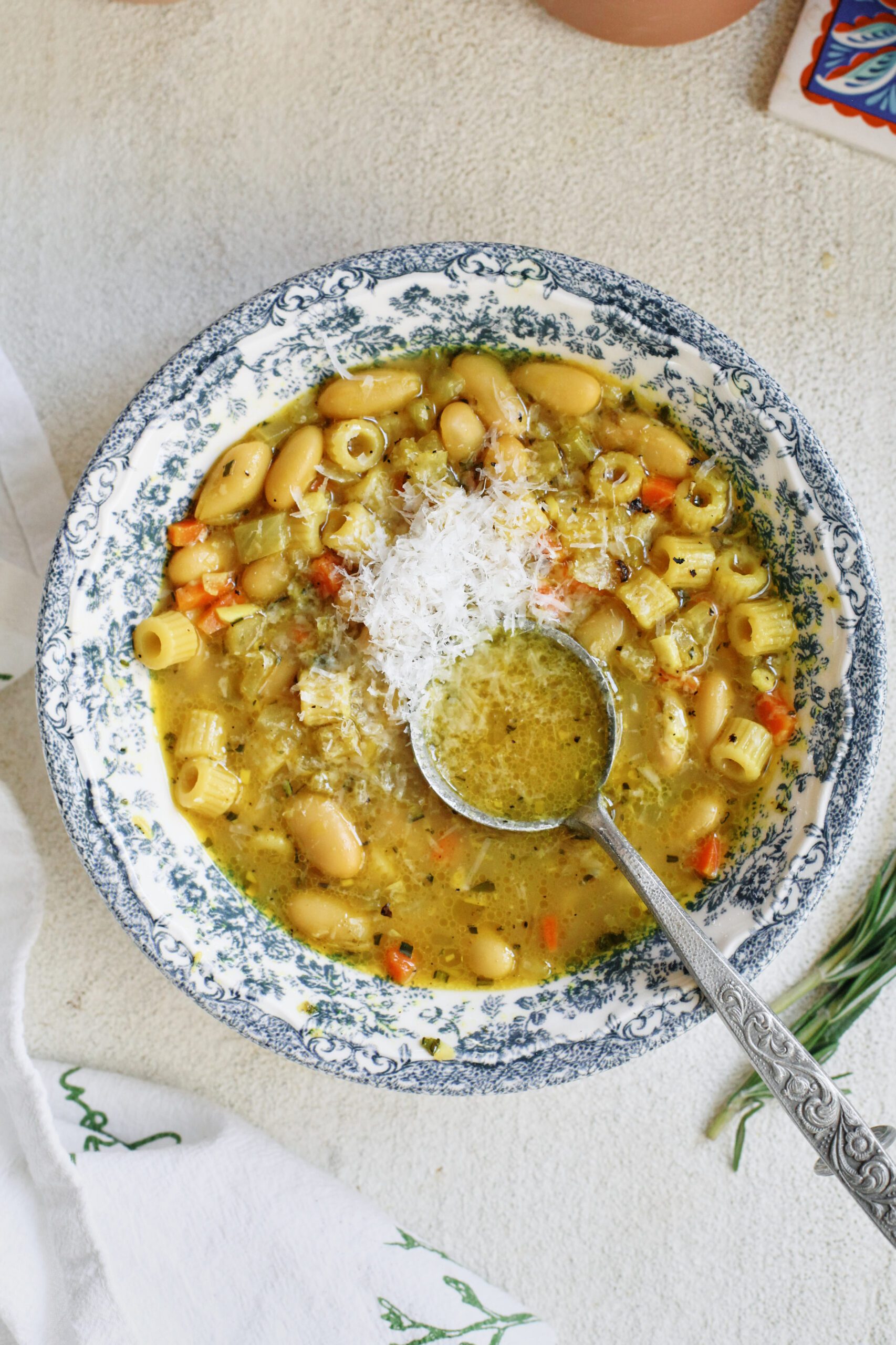 overhead photo of pasta e fagioli alla bianca in a blue/white floral patterned bowl. it's garnished with grated parmesan cheese and black pepper. an antique-looking silver spoon is in the bowl
