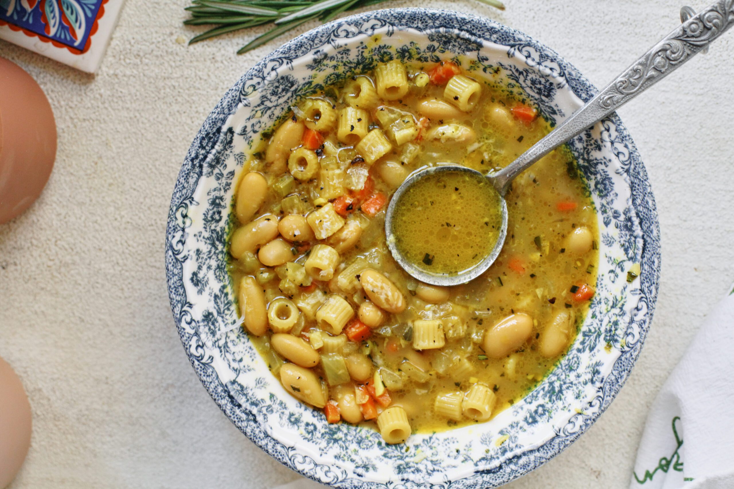 overhead photo of pasta e fagioli alla bianca in a blue/white floral patterned bowl. an antique-looking silver spoon is in the bowl