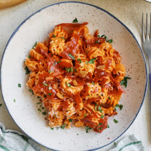 overhead photo of spicy vodka radiatore pasta with pieces crispy prosciutto and Italian parsley on top. It is in a white dish with a black rim, and theres a blue-handled silver fork next to the dish