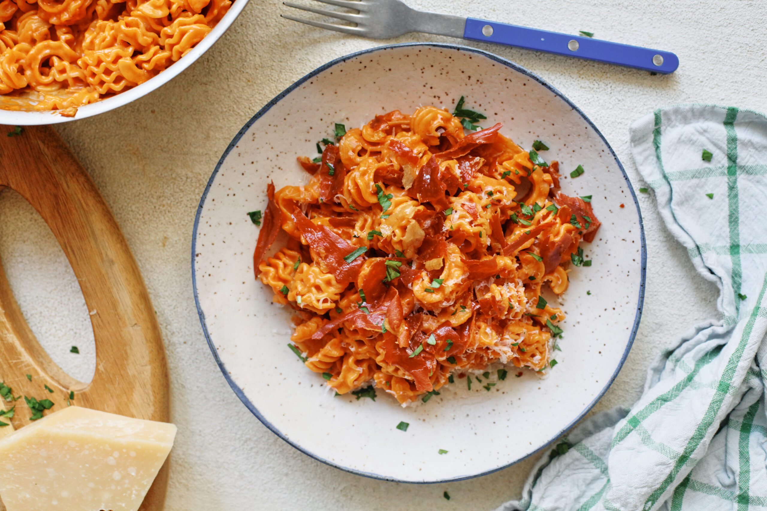overhead photo of spicy vodka radiatore pasta with pieces crispy prosciutto and Italian parsley on top. It is in a white dish with a black rim, and theres a blue-handled silver fork next to the dish