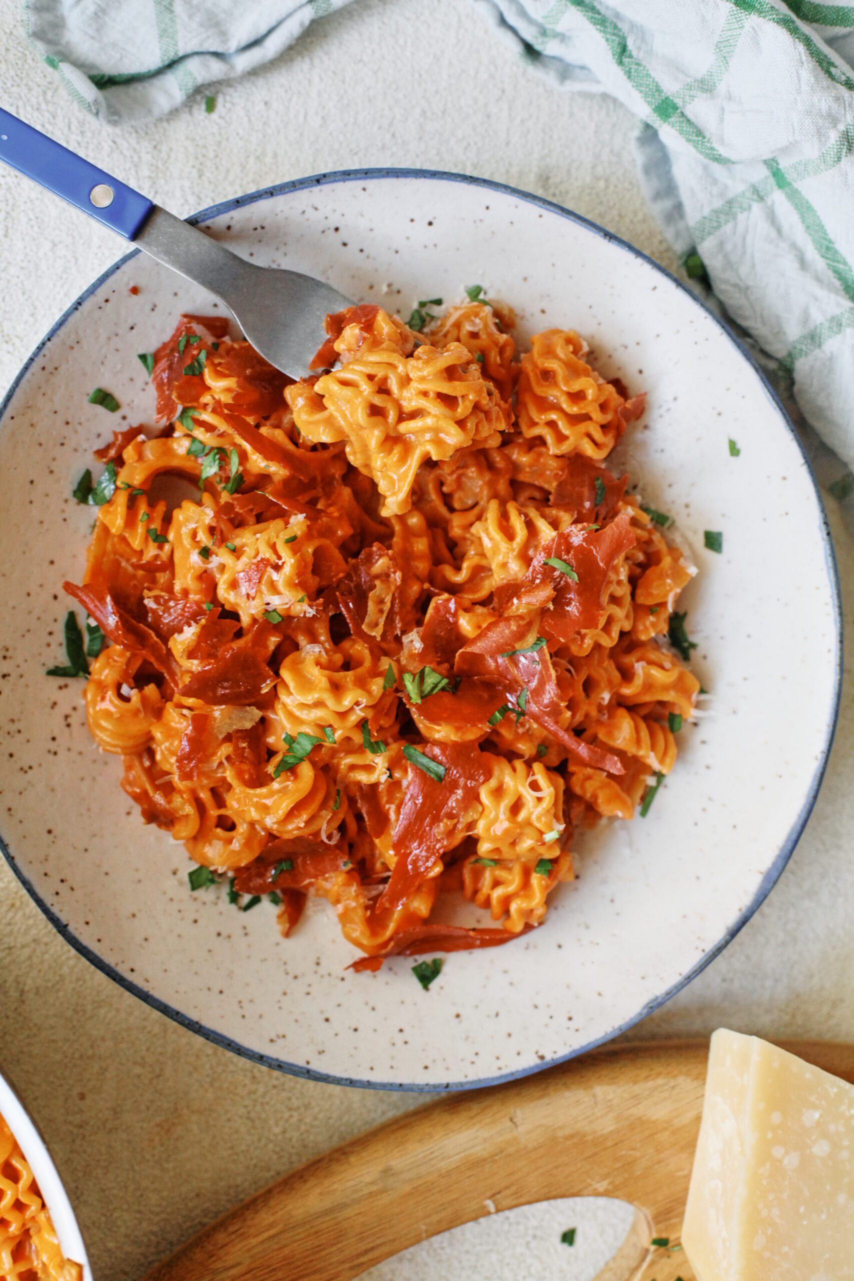 overhead photo of spicy vodka radiatore pasta with pieces crispy prosciutto and Italian parsley on top. It is in a white dish with a black rim, and theres a blue-handled silver fork in the dish