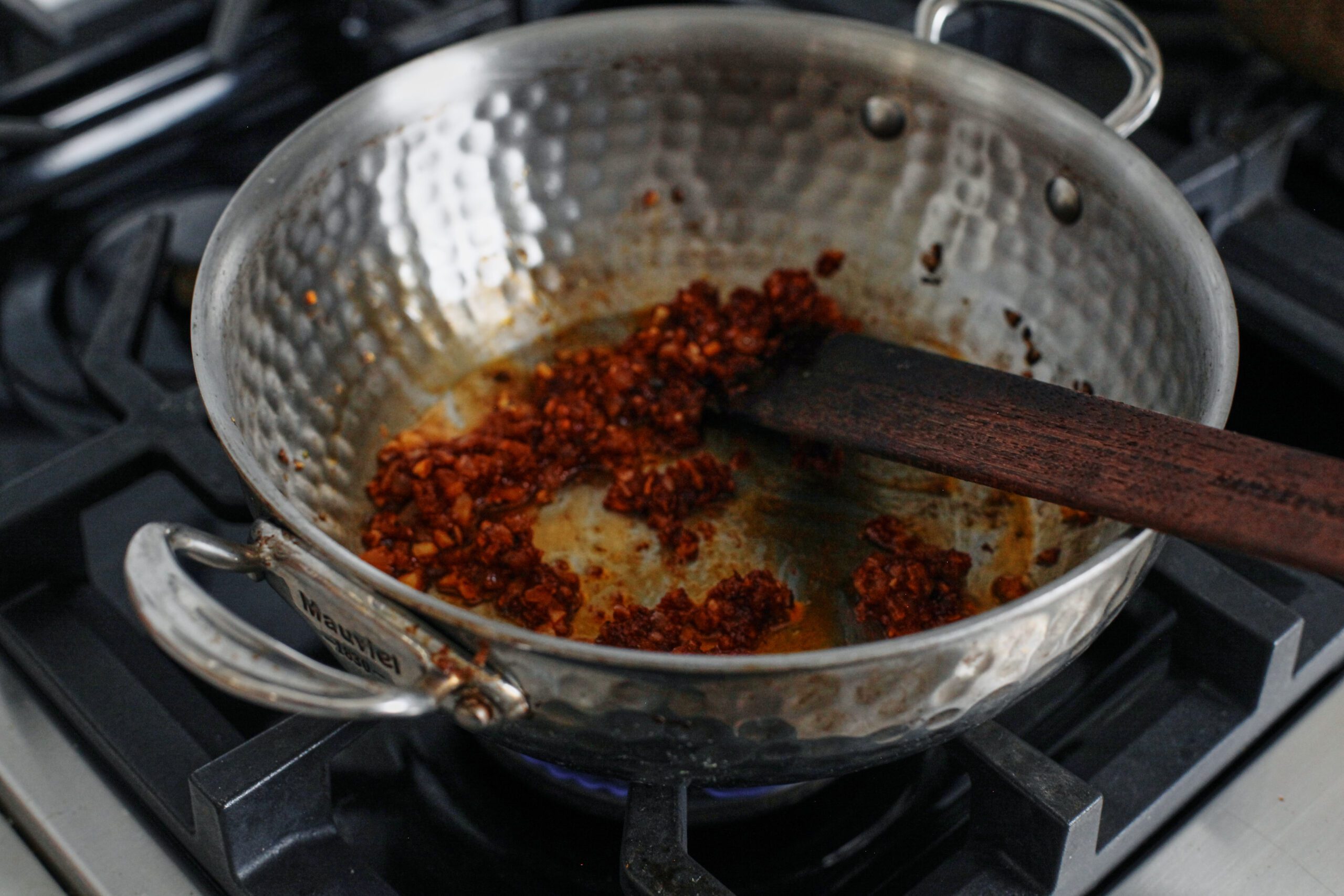 tomato paste being cooked with diced onion, minced garlic, and red pepper flakes in a hammered steel pan