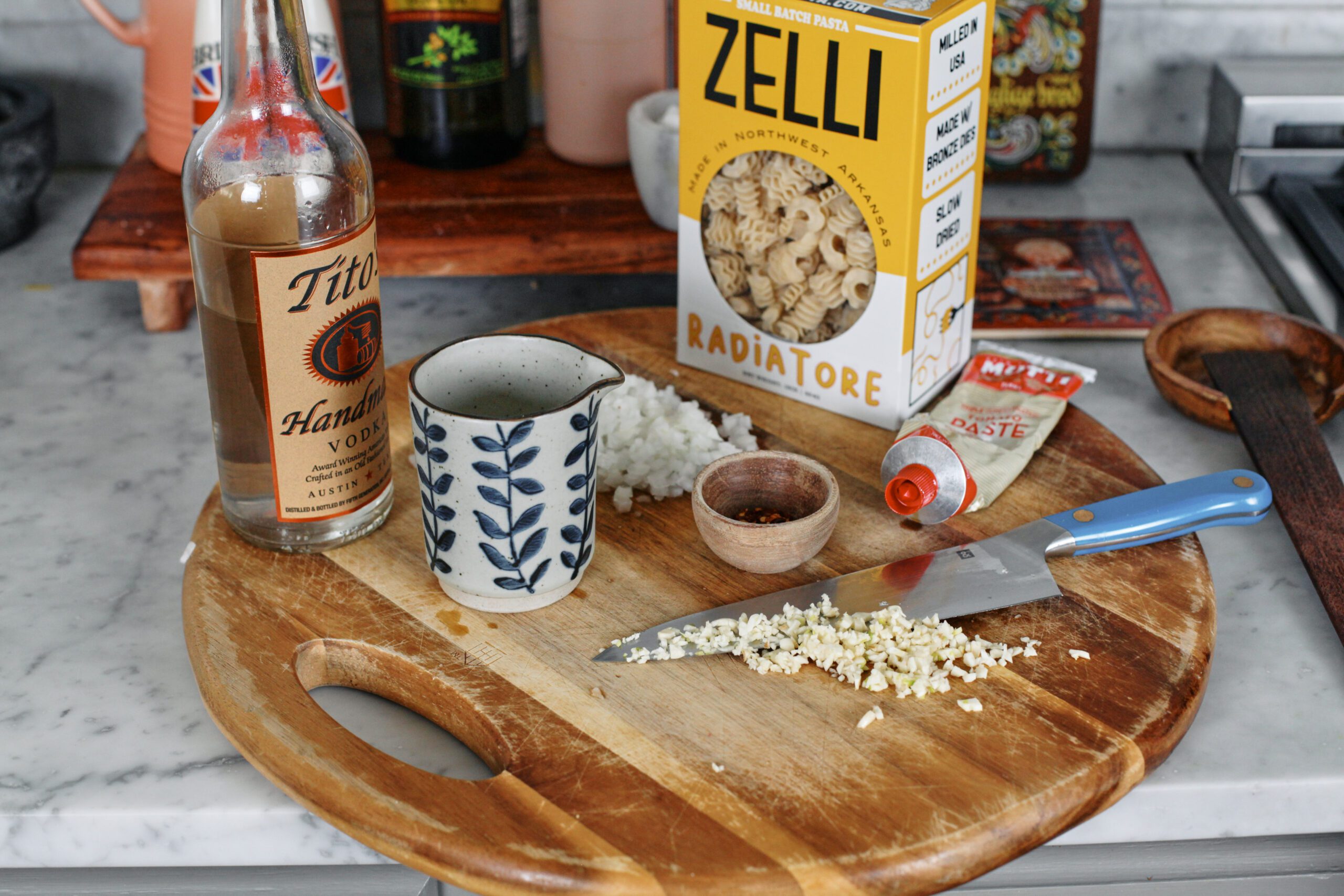 ingredients arranged on a round wooden cutting board: a bottle of Tito's vodka, a small carafe of heavy cream, 1/2 a yellow onion minced, a small bowl of red pepper flakes, minces garlic on the blade of a blue-handled chef's knife, a tube of tomato paste, and a box of radiatore pasta